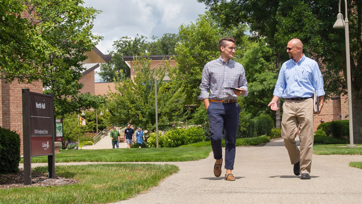 A student and a professor talk with each other as they walk on a campus path.
